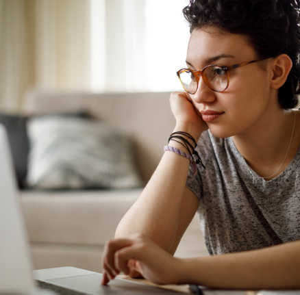 Image of a woman in glasses sat in front of a laptop, leaning up on her right arm and scrolling on her laptop with her left hand.