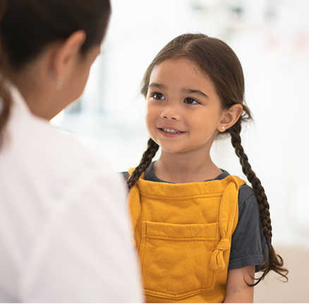 Image of a young girl with dark hair in 2 braids, wearing yellow dungarees, smiling and talking to a woman in a white shirt